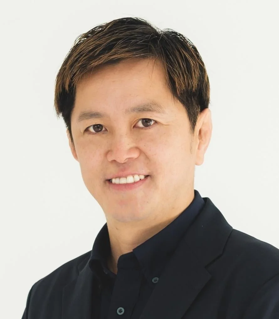 白い背景を背に、黒いシャツとジャケットを着用したアジア人男性の上半身ポートレート。男性は穏やかな笑顔を浮かべてカメラを見ている。ビジネスシーンやプロフィール写真に適した清潔感のある印象。