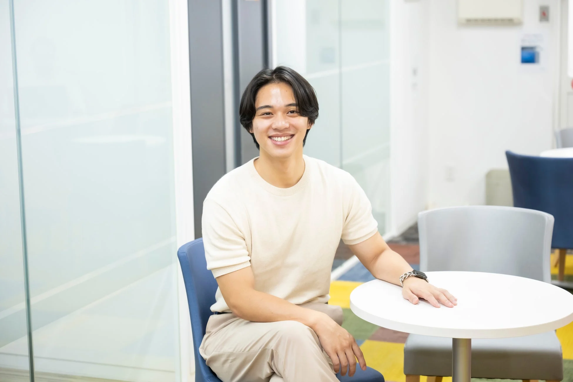 オフィスのような明るい空間で、ベージュの服を着た若い男性が笑顔で椅子に座り、テーブルに手をついている
