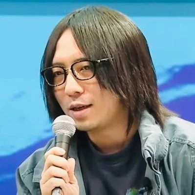 長髪でメガネをかけた男性がマイクを持ち、何かを話している様子