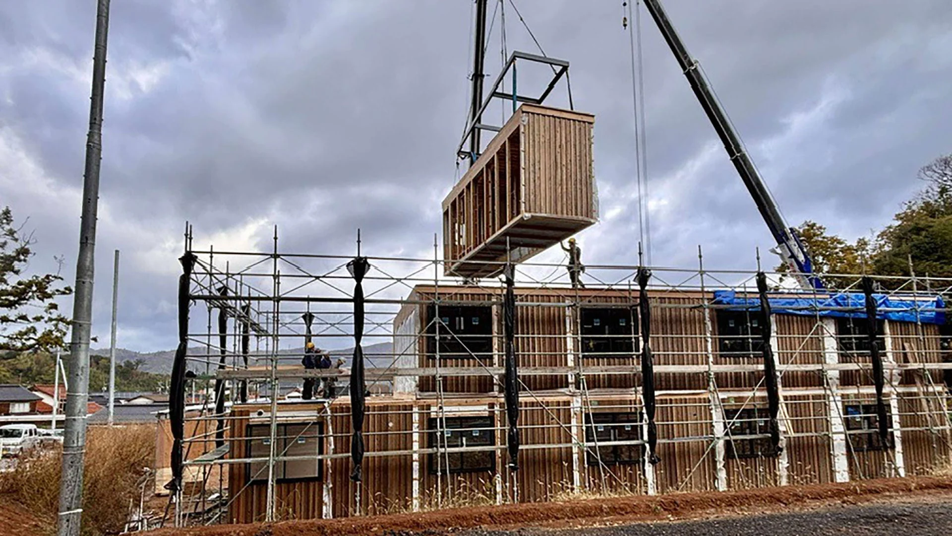 クレーンが木製のモジュール式ユニットを建設中の2階建ての建物に設置している様子。