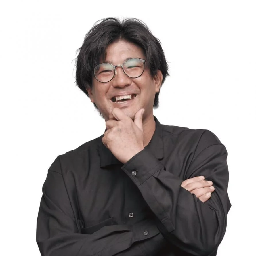 白背景の前で、黒いシャツと丸いメガネをかけたアジア人男性が、顎に手を当てて楽しそうに笑っています
