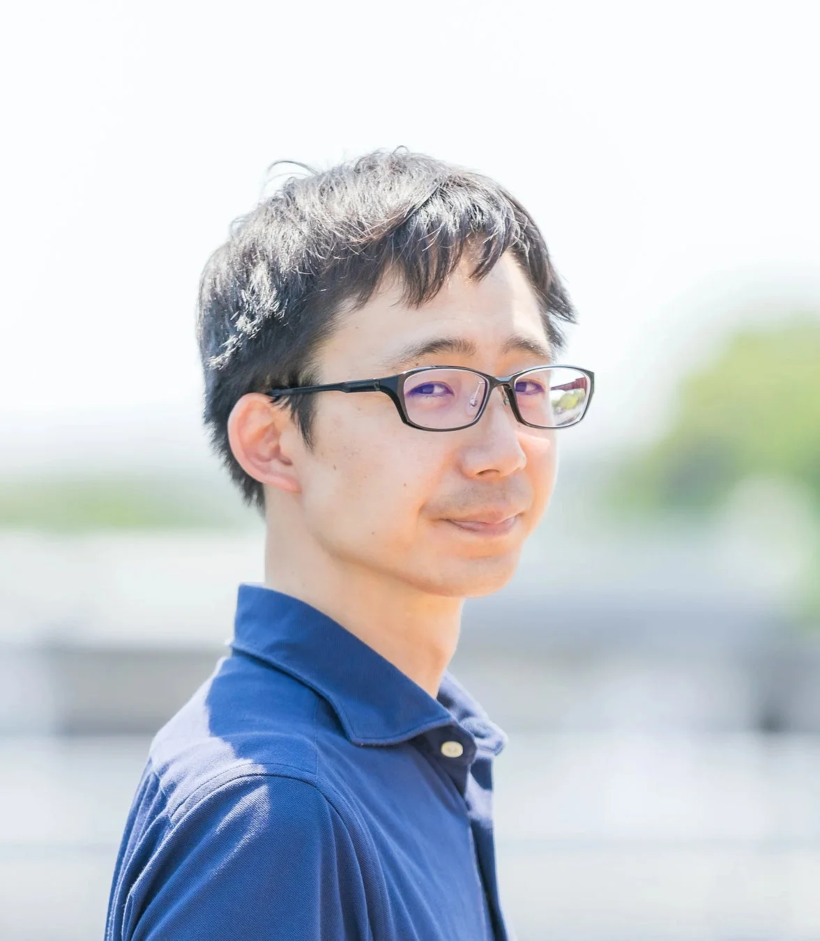 眼鏡をかけた男性が青いポロシャツを着て、明るい屋外で優しく微笑んでいるポートレート。