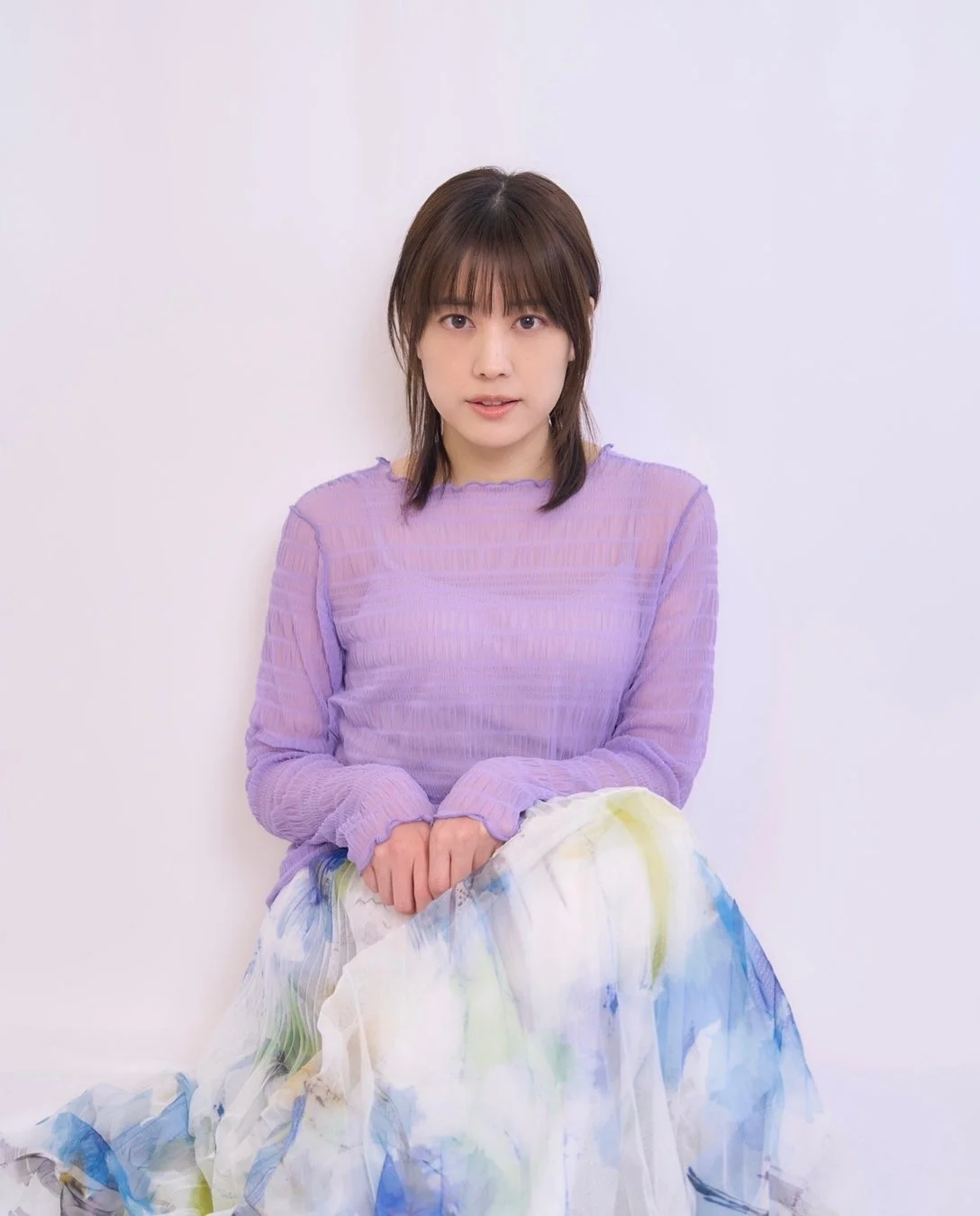 白い背景の前に座る女性のポートレート。透け感のある薄紫色のトップスと、水彩画のような模様の白いスカートを着用しており、カメラをまっすぐ見つめている。自然な表情が印象的。