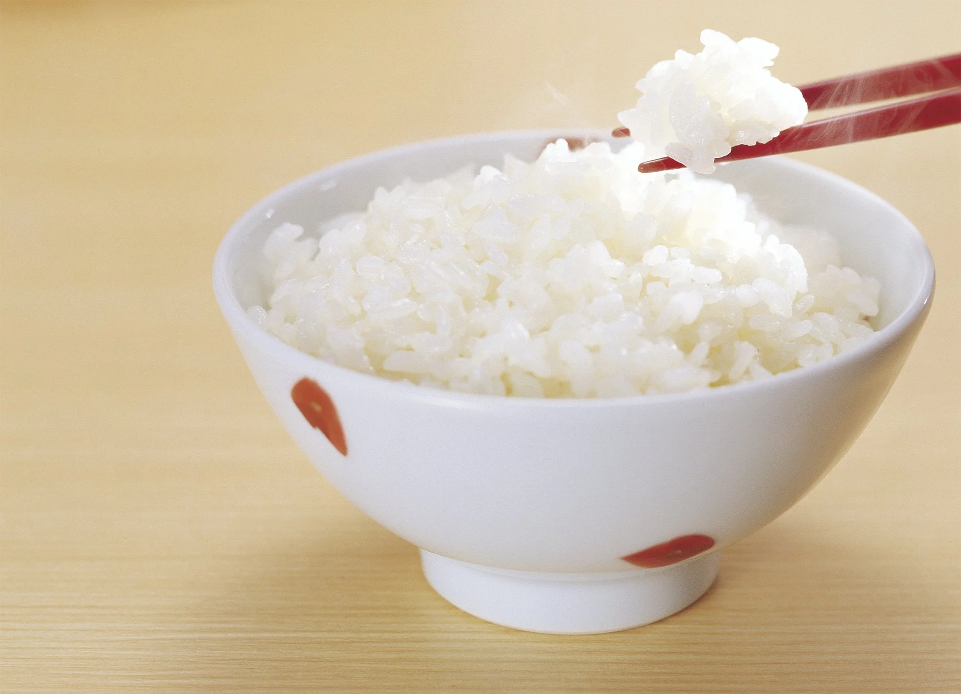 湯気の立つ炊きたての白いご飯が白い茶碗に盛られており、赤い箸でご飯をすくい上げている瞬間を捉えた写真です。温かさが伝わる食欲をそそる一枚。