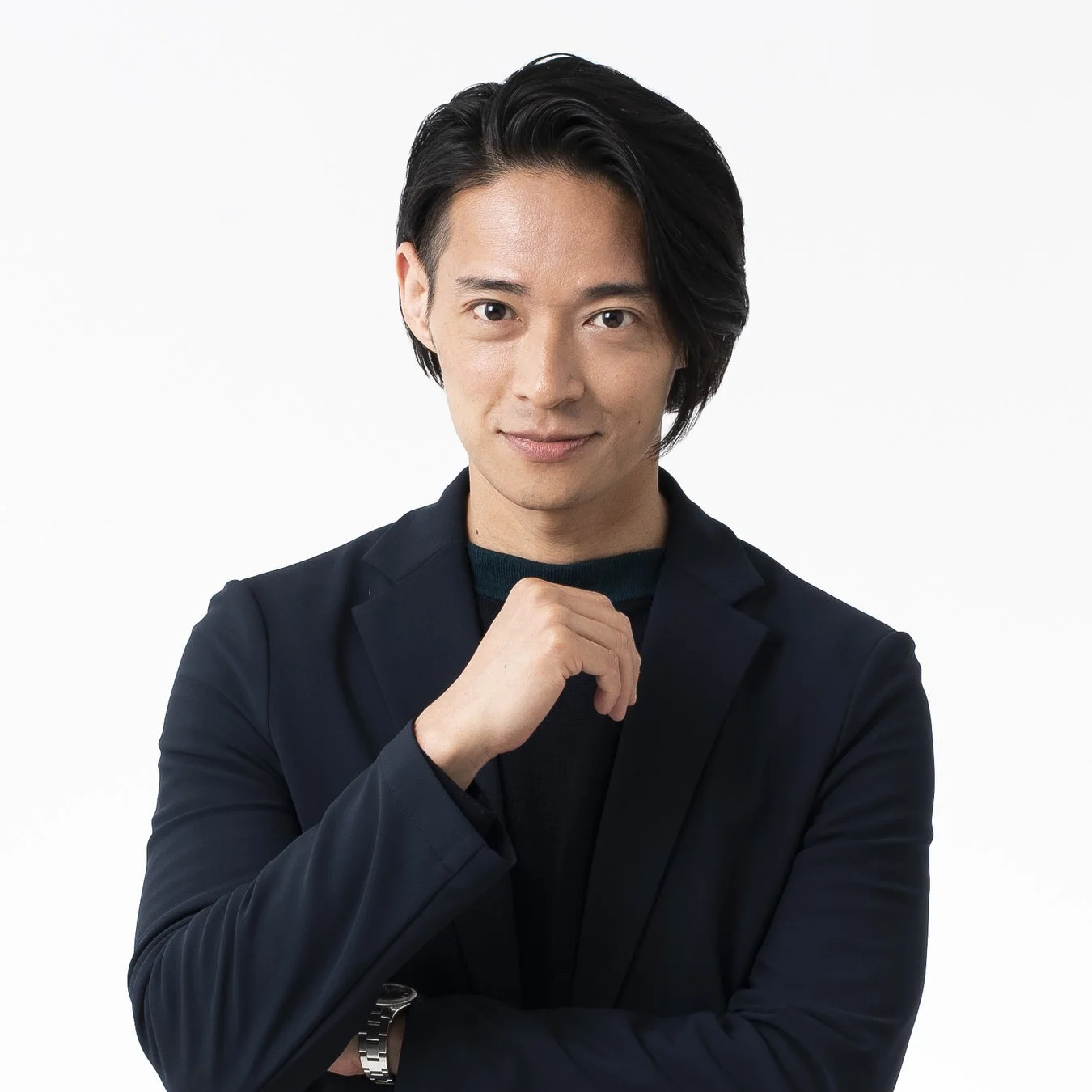 白背景の前で、ダークブルーのジャケットとダークグリーンのセーターを着た男性が、顎に手を当ててカメラを見つめているポートレート