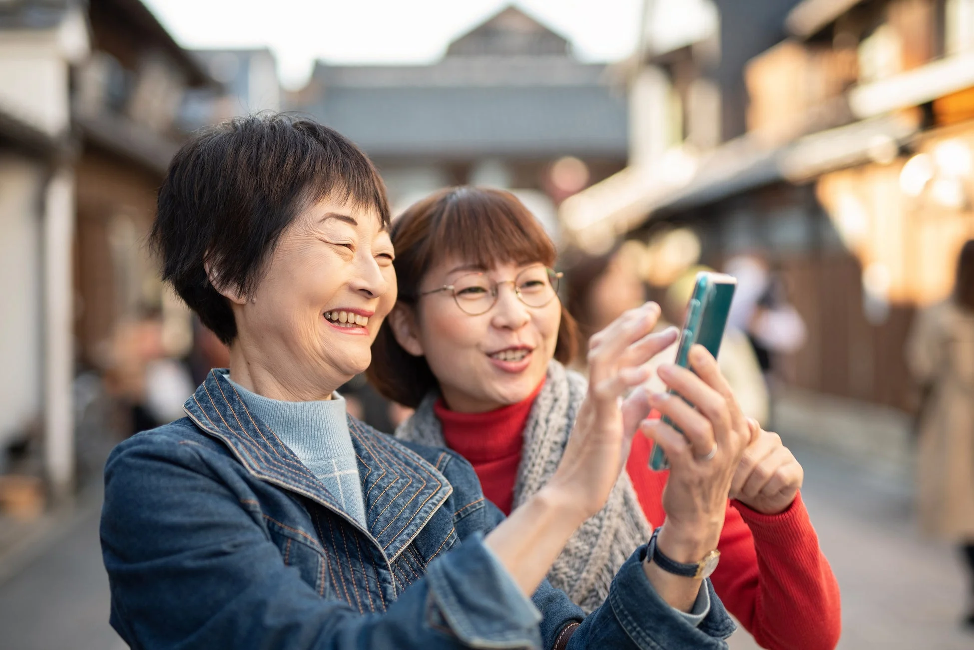 伝統的な街並みを背景に、二人のアジア人女性がスマートフォンを覗き込みながら満面の笑顔を見せている