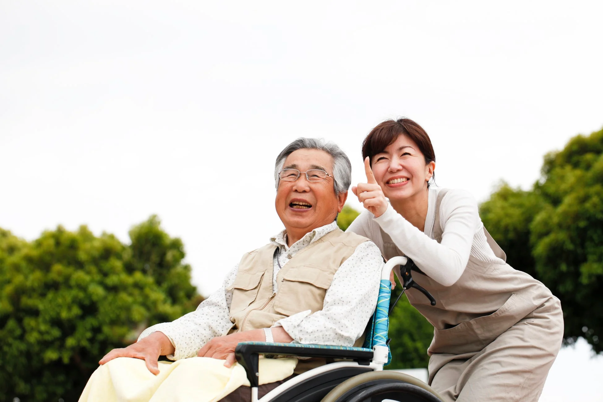 車椅子に乗った高齢男性と、彼を介助する女性が屋外で笑顔を見せている