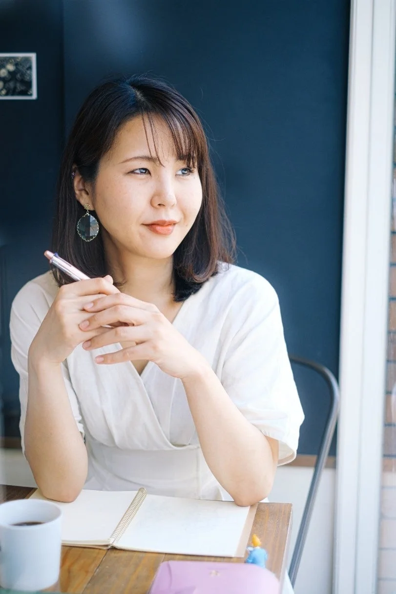 カフェの窓際に座る若い女性が、ペンを手に持ち、ノートを前にして窓の外を見つめています