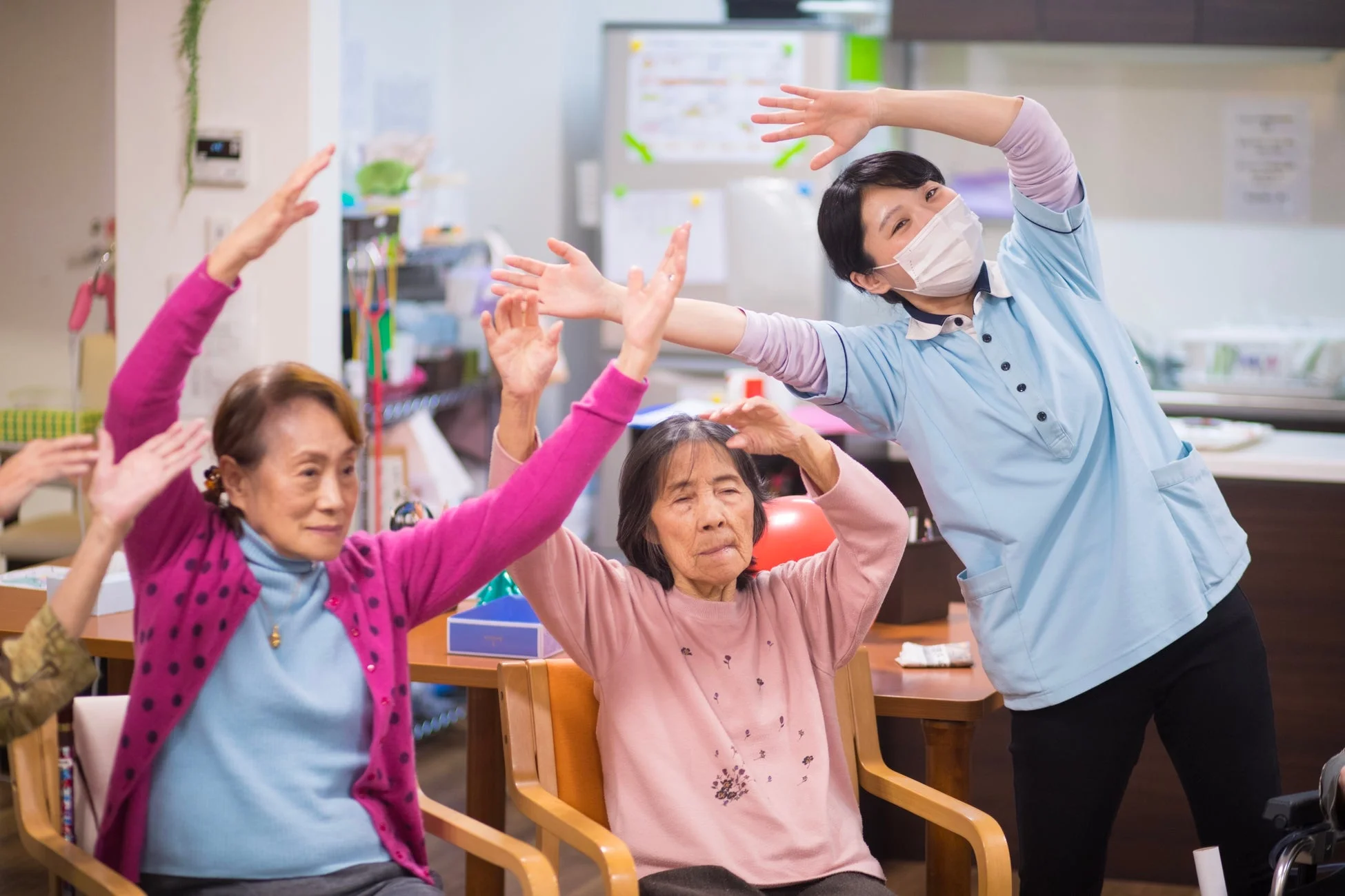介護施設で、介護士と高齢の女性2人が椅子に座って腕を上げる体操をしている