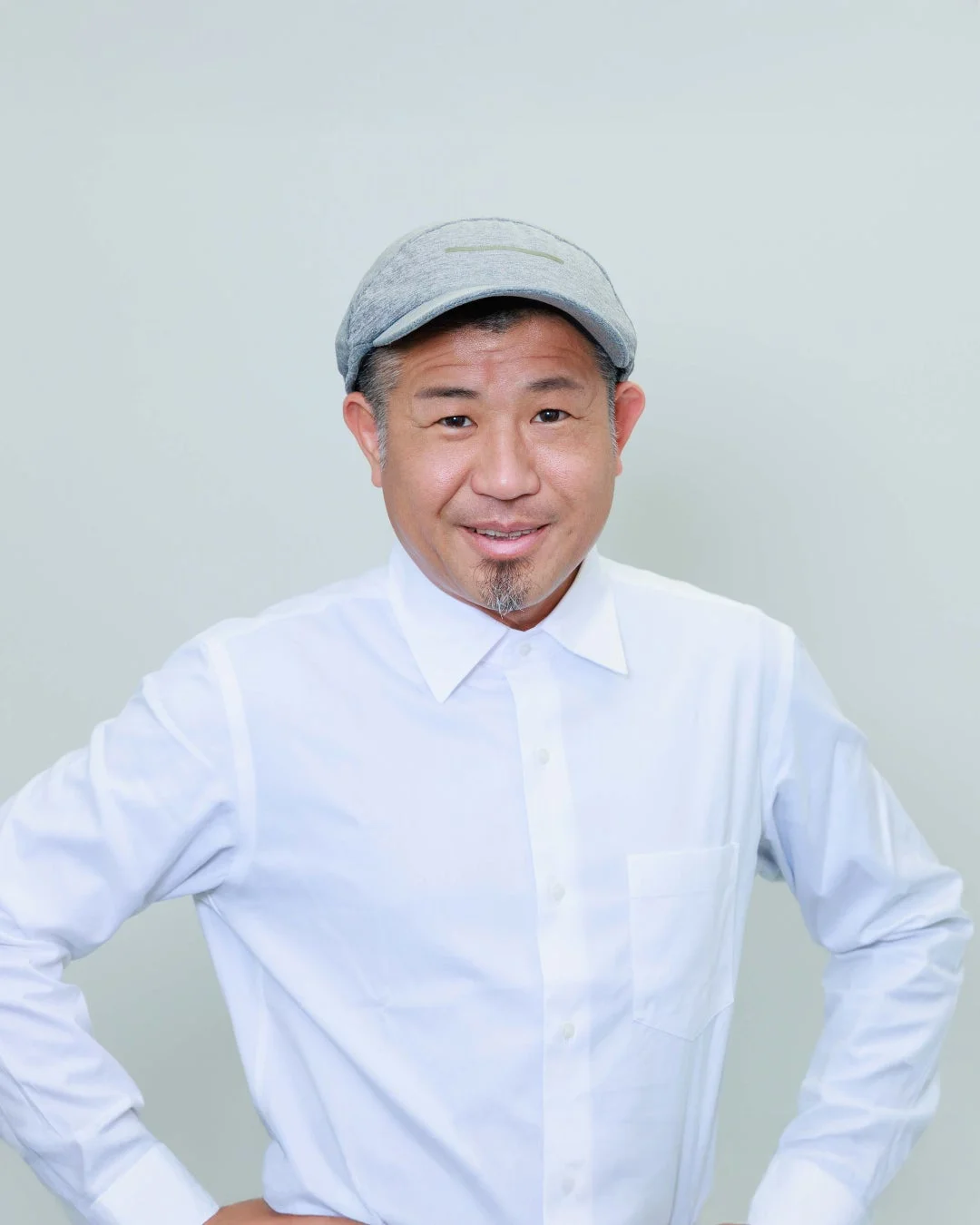 白いシャツとグレーの帽子を身につけた中年の男性が、白い背景の前で笑顔で立っているポートレート写真です。両手を腰に当てたポーズをとっています。
