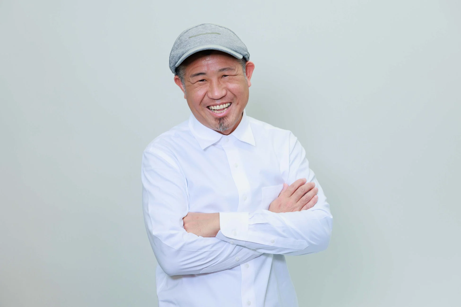 白いシャツとグレーの帽子を身につけた中年アジア人男性が、腕を組みながら明るい笑顔を見せているポートレート写真です。背景はシンプルな白です。