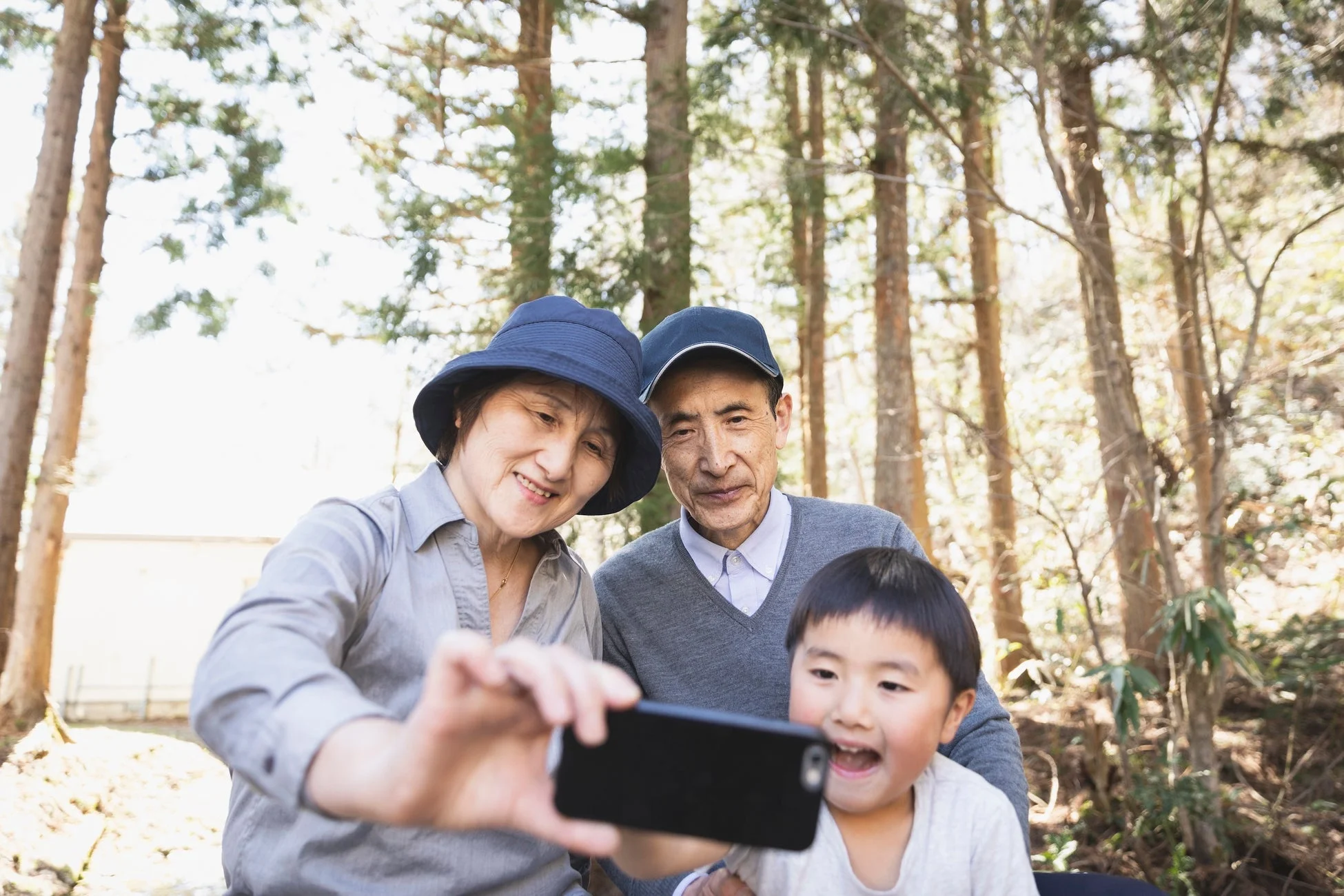 森林の中で、祖父母と孫がスマートフォンで楽しそうにセルフィーを撮っている様子