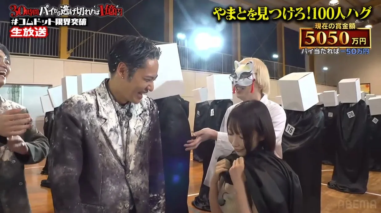 コムドットの生放送バラエティ番組「やまとを見つけろ!100人ハグ」のワンシーン。パイ投げゲームで参加者が白い粉まみれになっており、狐の仮面をつけた人物や箱を被った人々が映っている。現在の賞金額は5050万円と表示されている。