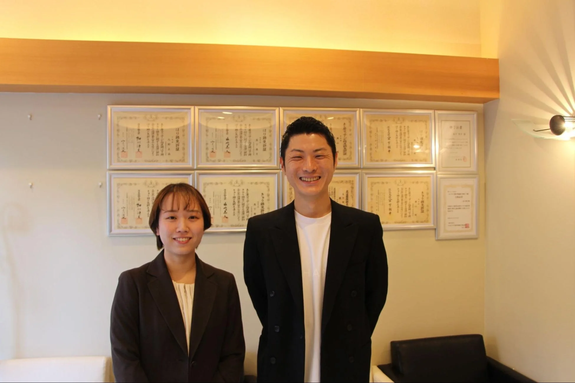 男女2人の専門家が、壁に飾られた鍼灸師やあん摩マッサージ指圧師などの複数の専門資格・免許証を背景に笑顔で立っている。信頼感とプロフェッショナリズムが伝わる一枚。