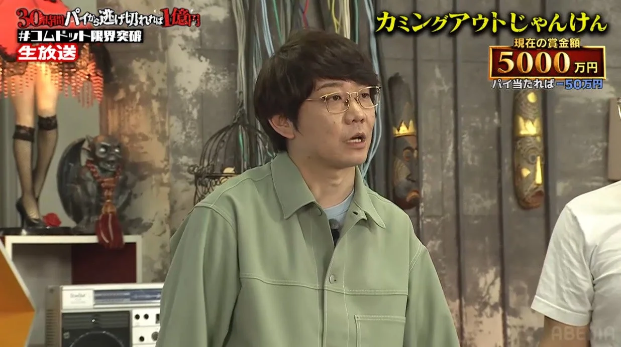 メガネをかけた男性がスタジオにいる様子です。画面には「30分間パイから逃げ切れば1億円」や「カミングアウトじゃんけん」といった番組に関するテロップがあり、現在の賞金額が5000万円であることが示されています。これは「コムドット 限界突破」という生放送のゲーム番組の一場面のようです。