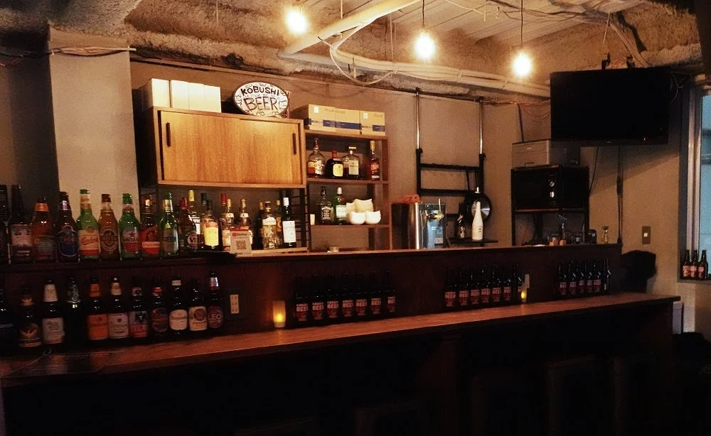 薄暗いバーの店内。長い木製カウンターには、様々な種類のビールや洋酒のボトルが並べられている
