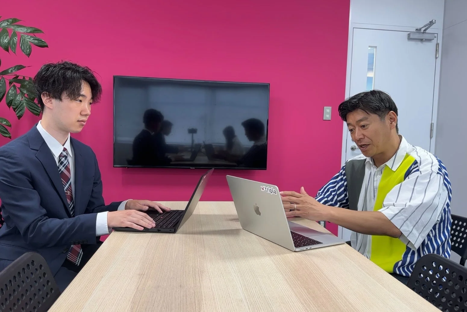 ピンクの壁を背景に、2人の男性がノートパソコンを前にテーブルに向かい合って座っています。
